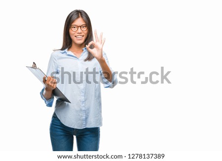 Young asian business woman over isolated background holding clipboard doing ok sign with fingers, excellent symbol