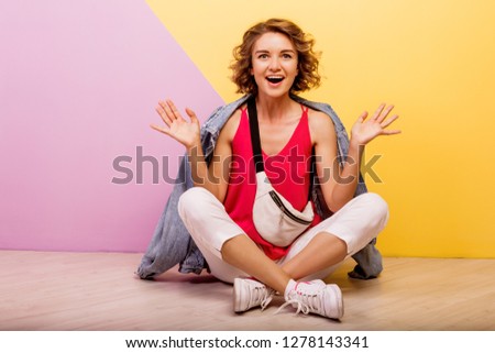 Hipster  woman with surprise face   posing in studio on pink and yellow background. Happy dreamy mood. Wearing summer sporty outfit, sneakers and jeans jacket. 