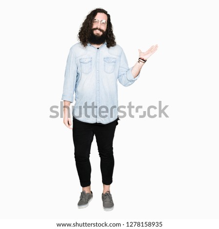 Young hipster man with long hair and beard wearing glasses smiling cheerful presenting and pointing with palm of hand looking at the camera.