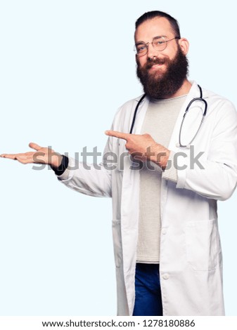 Young blond doctor man with beard wearing medical coat amazed and smiling to the camera while presenting with hand and pointing with finger.