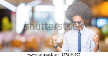 Young african american business man with afro hair wearing sunglasses very happy and excited doing winner gesture with arms raised, smiling and screaming for success. Celebration concept.