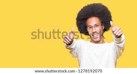 Young african american man with afro hair wearing glasses approving doing positive gesture with hand, thumbs up smiling and happy for success. Looking at the camera, winner gesture.