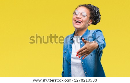 Young braided hair african american girl wearing glasses over isolated background smiling friendly offering handshake as greeting and welcoming. Successful business.