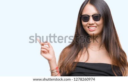 Young asian woman wearing sunglasses over isolated background with a big smile on face, pointing with hand and finger to the side looking at the camera.