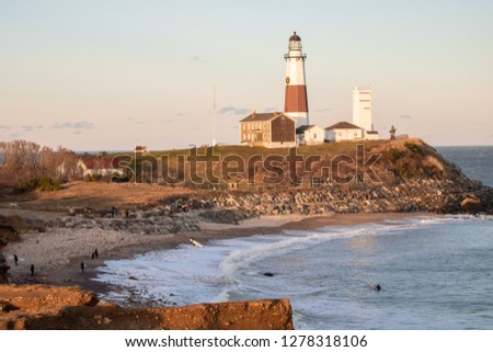 Surfers catching waves on a cold afternoon as golden light washes over a nearby lighthouse. Montauk State Park, New York