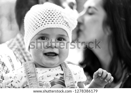 Girl cute smiling baby hazel-eyed kid tiny little child wearing white flower beanie hat in arms of kissing parents mother and father family summer day on blurred green background, horizontal picture 