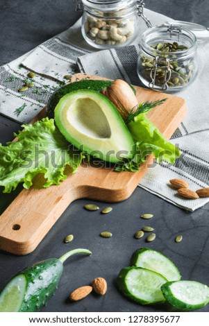 Still life, sliced avocado lies on a piece of lettuce on a wooden board on a cotton napkin. Nearby is sliced cucumber, almonds and pumpkin seeds. In the background are glass jars with nuts.