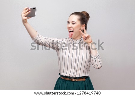 Selfie time! Portrait of happy foolish joyful attractive blogger woman wearing in striped shirt standing, winking and showing tongue and making selfie. Indoor, isolated, studio shot, grey background