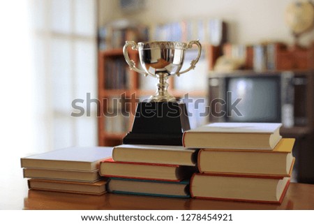 Close-up pictures of many books stacked on the table in which there are trophies on top. Bookshelf in the background selective focus and shallow depth of field