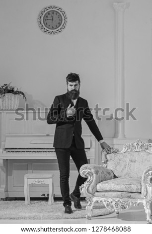 Man with beard and mustache wearing classic suit, stylish fashionable outfit. Macho attractive and elegant on serious, thoughtful face standing near old fashioned armchair. Fashion and style concept.