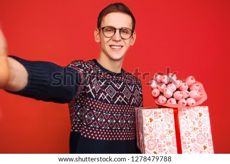 A man in glasses, with a gift in his hands and a bouquet of flowers, photographs himself on a smartphone against a red background. Valentine's Day