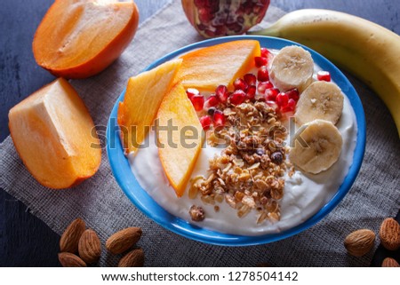 A blue plate with greek yogurt, granola, persimmon, banana, pomegranate black wooden background. close up.
