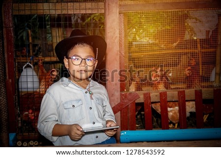 Portrait asian boy wears glasses, 7 years old, wearing a cowboy outfit, holding a tablet, playing on a chicken farm for education.
