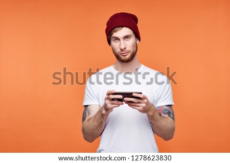 A man in a red cap with tattoos on his hands and in a white t-shirt is holding a phone on an orange background               