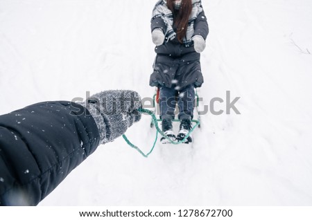 Winter season, snowing outside. Riding the sledge concept. The man is pulling the colored sled behind himself with a woman on it. Hand in glove with a sled rope.