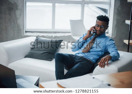 Break time. Photo of afro american young guy sitting on a couch and have conversation at phone.