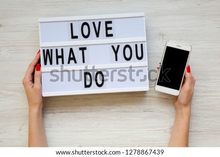 Female hands hold modern board with text 'Love what you do' and smartphone over white wooden background, top view. Business concept. From above, flat-lay, overhead. 