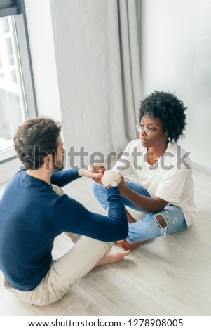 Dark skinned woman and caucasian bearded handsome man sit on floor together in lotus pose in empty room, practice yoga near the big window.