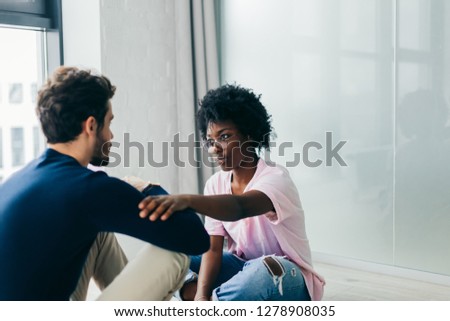 Caucasian boyfriend and his african girlfriend practice yoga, sit crossed legs, holding hands together, looking at each other while sitting on wooden floor in spacious room.