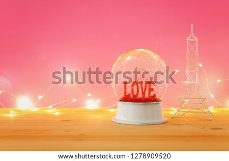 Valentine's day background. Water globe with word LOVE and glitter next eiffel tower, over the wooden table and pink bakground