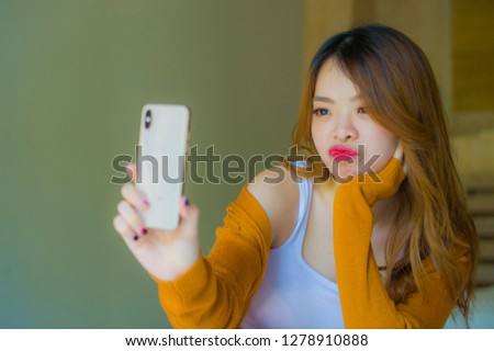 young attractive and relaxed Asian Chinese girl at home bedroom enjoying taking selfie portrait photo with mobile phone camera sitting on bed doing faces in teenager student lifestyle