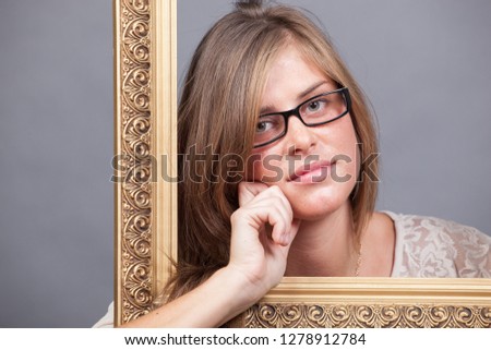 young woman in eyeglasses looking through wooden golden frame on grey background