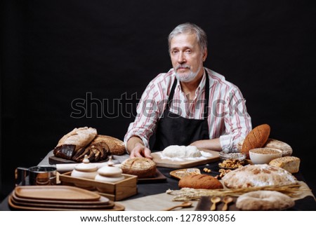 Elderly professional chef man wears apron as being on kitchen, tired after baking and cooking, looking at camera, while sitting at table with ingridients for preparing bread and dough at table
