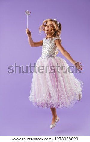 Full length portrait of a smiling pretty girl dressed in a princess dress isolated over violet background, holding magic wand, jumping