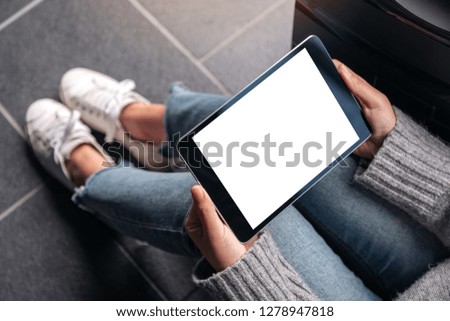 Top view mockup image of woman's hands holding and using black tablet pc with blank white desktop screen while sitting on the floor 