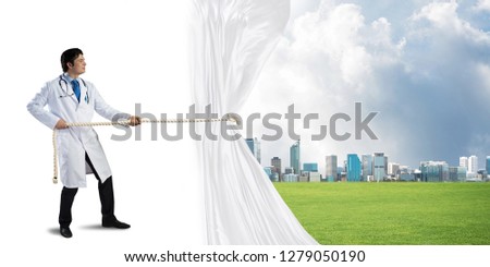 Horizontal shot of young doctor in white medical uniform pulling white fabric while standing over white background with cityscape view behind curtain