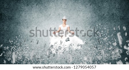 Woman in white clothing keeping eyes closed and looking concentrated while meditating on flying cloud among flying letters with gray dark wall on background.