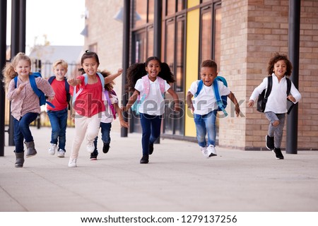 A group of smiling multi-ethnic school kids running in a walkway outside their infant school building after a lesson Royalty-Free Stock Photo #1279137256