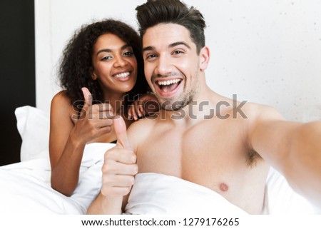 Beautiful happy young multiethnic couple relaxing in bed under blanket, taking a selfie