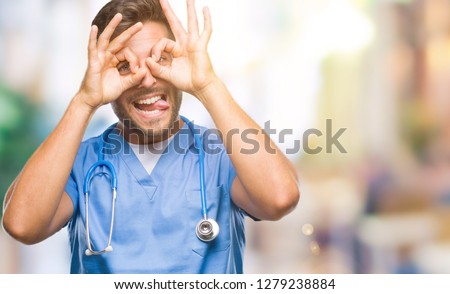 Young handsome doctor surgeon man over isolated background doing ok gesture like binoculars sticking tongue out, eyes looking through fingers. Crazy expression.