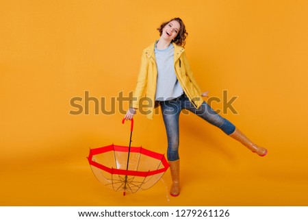 Amazing girl in blue jeans and shirt funny dancing, holding umbrella and smiling to camera. Studio shot of joyful lady in autumn jacket and rubber shoes having fun after rain.