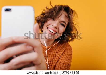 Close-up portrait of adorable young woman with wavy hair making selfie and laughing. Indoor photo of stunning brunette girl in knitted sweater taking picture of herself.