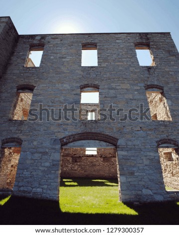 Tan stone wall of an abandoned historical building which towers over the viewer. The home is in ruins and you can see the bright light shine through the empty windows and onto bright green grass.