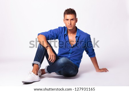 full length picture of a serious young casual man sitting on the floor and looking into the camera, on light background