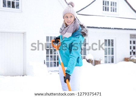 Pretty young blonde woman in winter sheds the white snow in front of her house on a big pile - dressed in cold weather blue jacket and cap she smiles happily