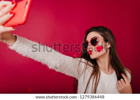 Enchanting woman with heart on cheek posing with kissing face expression. Indoor photo of charming girl in funny glasses using smartphone for selfie.