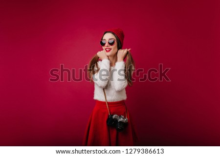 Enchanting white woman with camera posing with dreamy face expression. Indoor photo of charming european female photographer wearing red skirt.