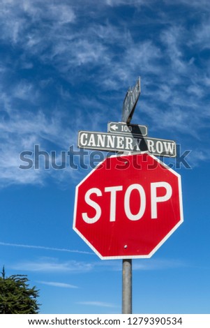 Looking up at a bold red octagonal STOP sign contrasted against a bright blue sky with light cloud. The sign marks the junction to Cannery Row and Reeside Avenue, Monterey, California, USA.