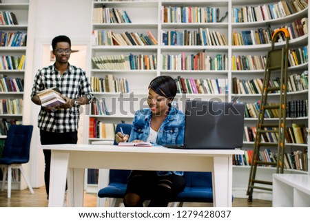 Pretty young african girl in casual clothes writing and studying with notebook and books in university library. Handsome african man in glasses holding books going to the girl on the background.