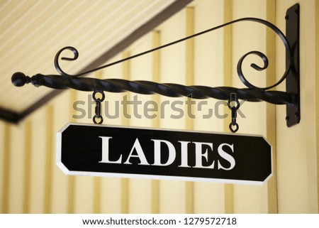 A Ladies sign hanging from a metal rod with a scroll on top over a bathroom.