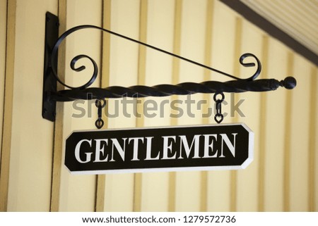 A Gentlemen sign hanging from a metal rod with a scroll on top over a bathroom.