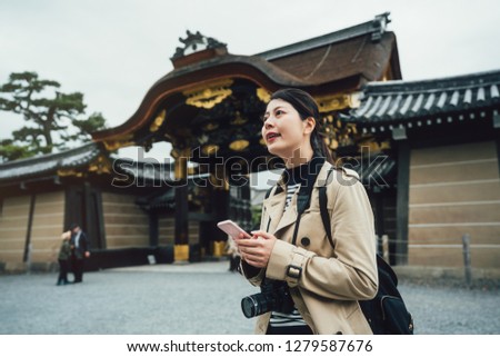 Cheerful asian female traveler holding cellphone searching direction online map app. young girl photographer carrying camera standing near nijo jo gate on sunny day in spring vacation kyoto japan.