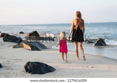 Mother in black dress and daughter in pink dress walk hand in hand on the beach. Blondies on the ocean