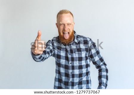 Red hair mature man standing isolated on grey wall showing thumb up tongue out grimacing playful to camera