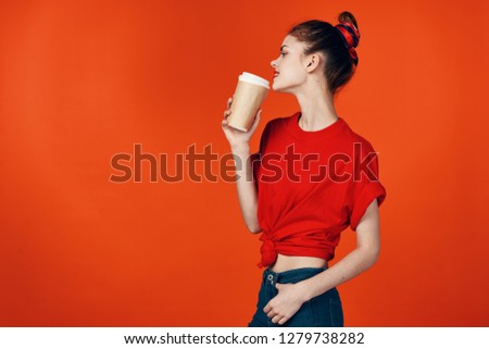 A young woman in a red t-shirt and jeans holds a glass of coffee in her hand.                