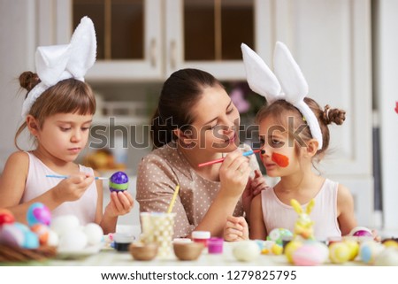 Mother draws on daughter's nose while she and her two little daughters with white rabbit's ears on their heads dye the eggs for the Easter table in the cozy light kitchen.